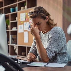 Exhausted businesswoman having a headache at office. Mature creative woman working at office desk feeling tired. Stressed casual business woman feeling eye pain while overworking on desktop computer.