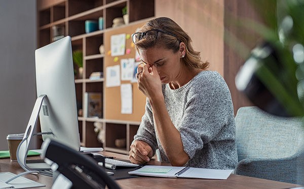 Exhausted businesswoman having a headache at office. Mature creative woman working at office desk feeling tired. Stressed casual business woman feeling eye pain while overworking on desktop computer.